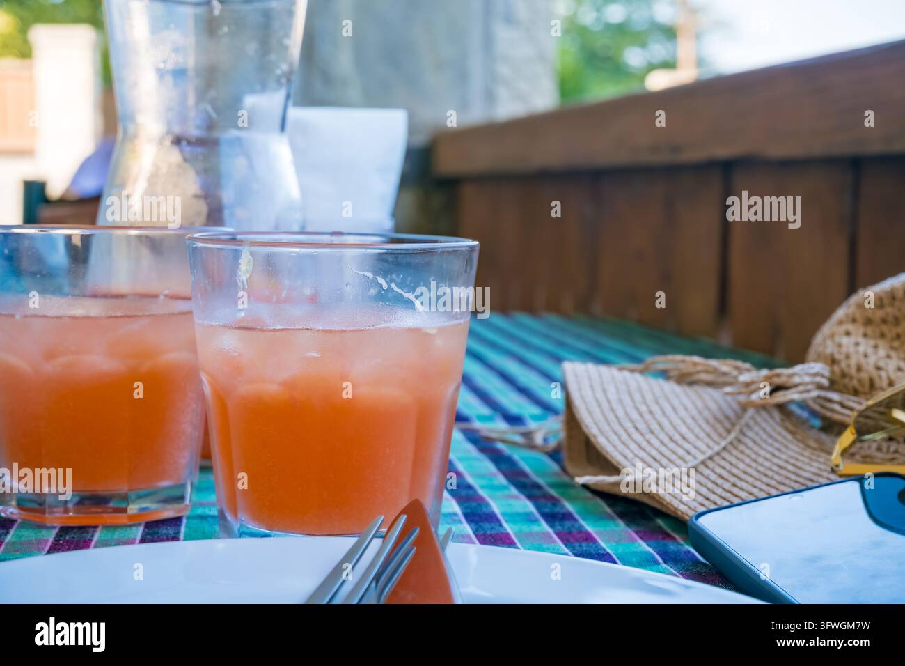 Due bicchieri raffreddati di limonata rosa su un tavolo all'aperto colorato con una brocca di vetro, piatti, forchette, tovagliolo piegato, occhiali da sole e panca di legno Foto Stock