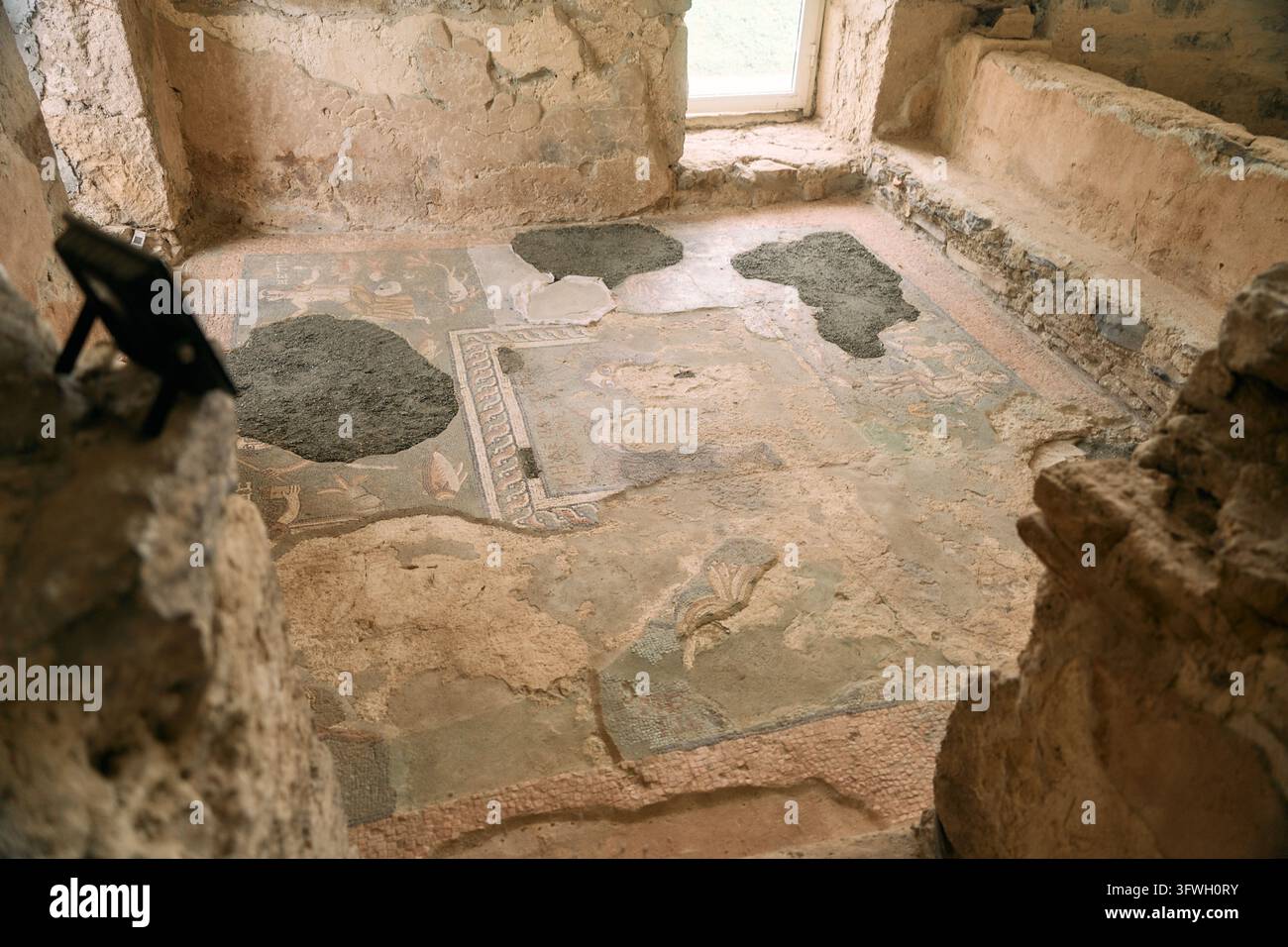 Mosaico di pavimento antico consumato all'interno di una rovina archeologica con malta rattoppata e muri di pietra che si sgretolano sotto una luce soffusa della finestra, conservazione del patrimonio Foto Stock