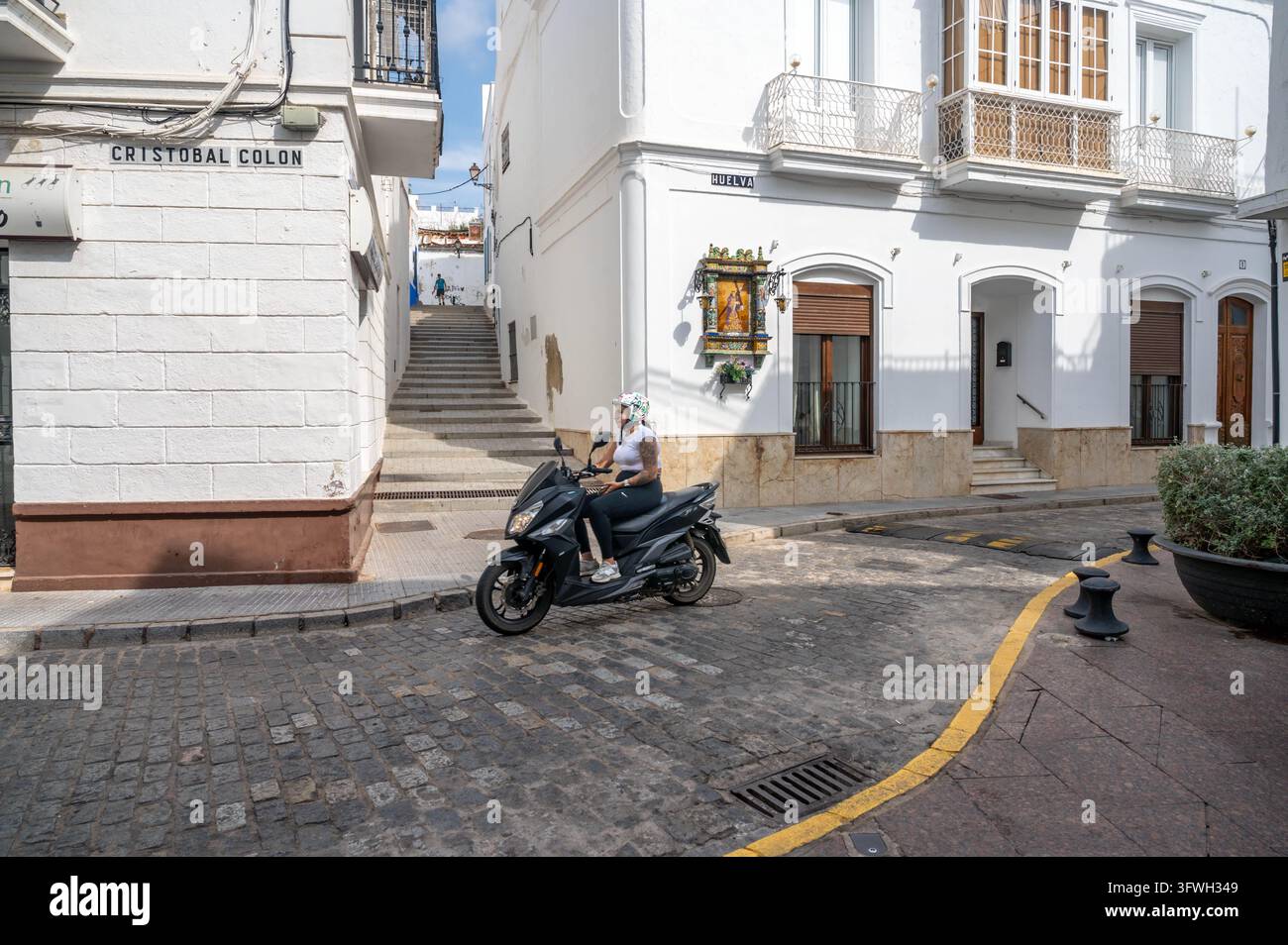 Ayamonte, Spagna, 10-03-2026: Donna su uno scooter elettrico guida su una vecchia strada acciottolata nella città spagnola di Ayamonte, passaggio pedonale e chiesa Foto Stock