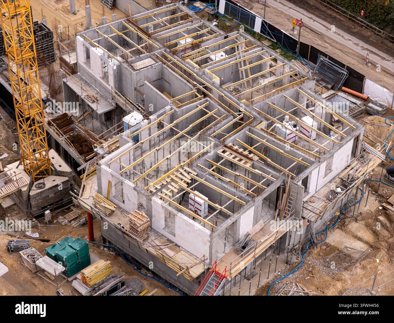 Vista aerea di un cantiere edile con una struttura in cemento con travi del tetto in legno, una gru a torre gialla e vari materiali da costruzione in Foto Stock