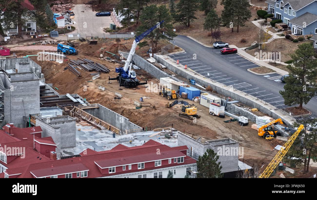 Veduta aerea di un cantiere edile con una gru blu, un escavatore giallo e mezzi pesanti vicino a un grande edificio con tetto rosso a Estes Park Foto Stock