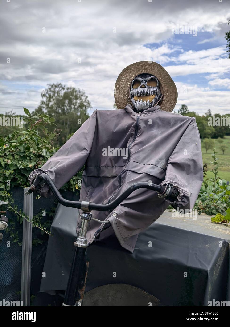 Figura di spaventapasseri in bicicletta nel giardino con maschera da teschio e cappello di paglia sotto le nuvole Foto Stock