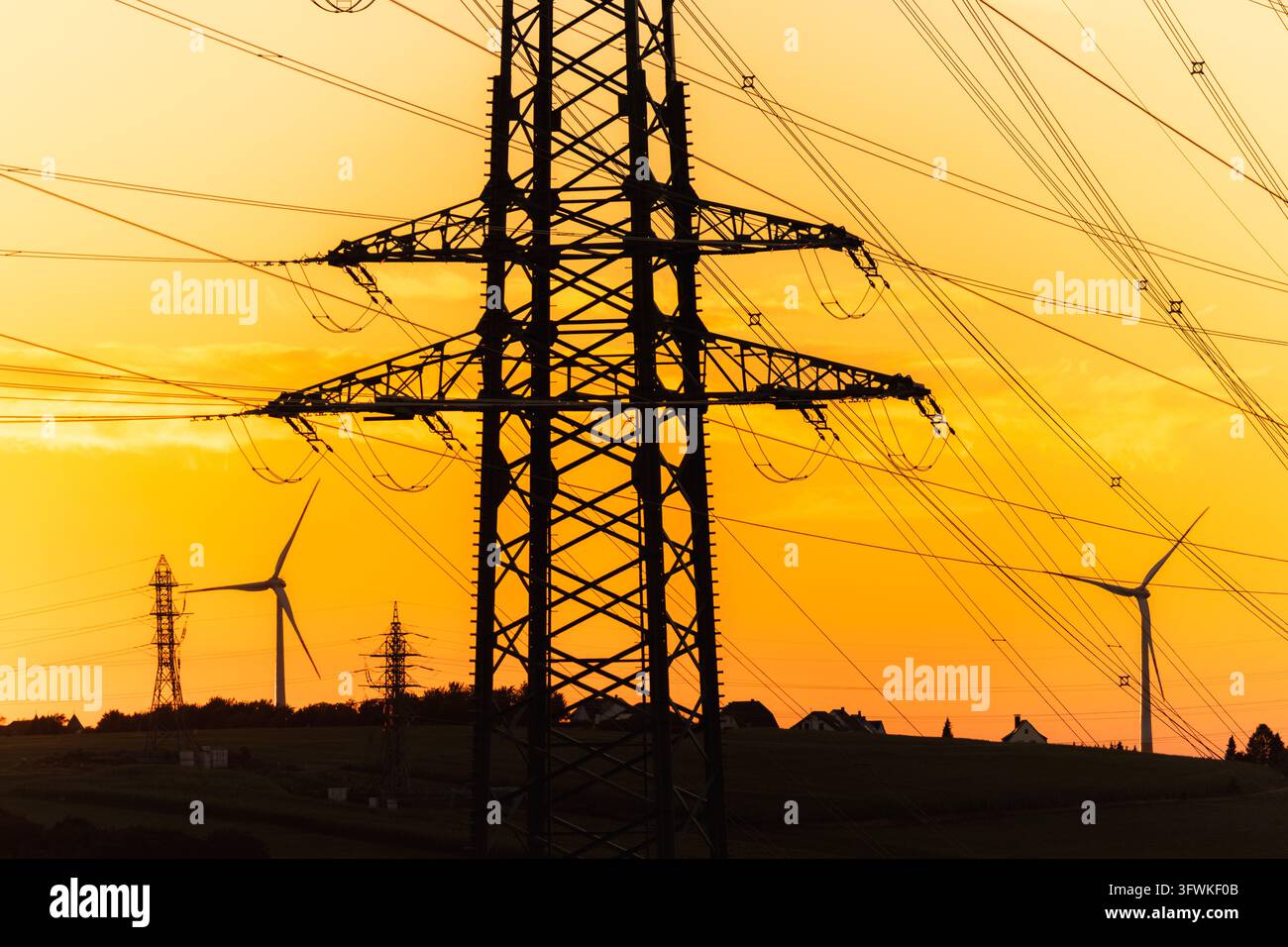 Torri di trasmissione di energia ed eoliani formano un paesaggio energetico in silhouette sotto un cielo luminoso al tramonto. Foto Stock