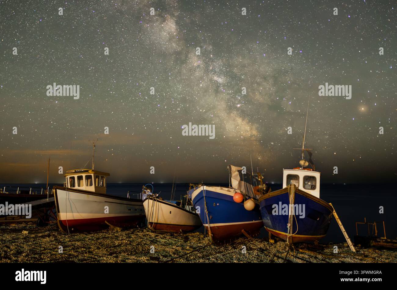 Inghilterra, Devon, Beer. Un campo di stelle fitto e un nucleo galattico luminoso si ergono sopra barche da pesca ormeggiate, con le luci calde del porto che illuminano la riva rocciosa Foto Stock