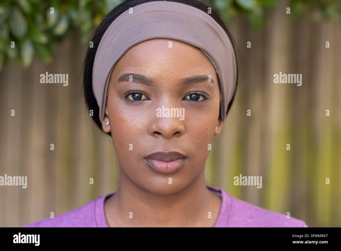 Donna adulta africana in posa nel cortile vicino a una recinzione di legno, indossa una fascia beige, una maglia a collo alto malva e borchie Foto Stock