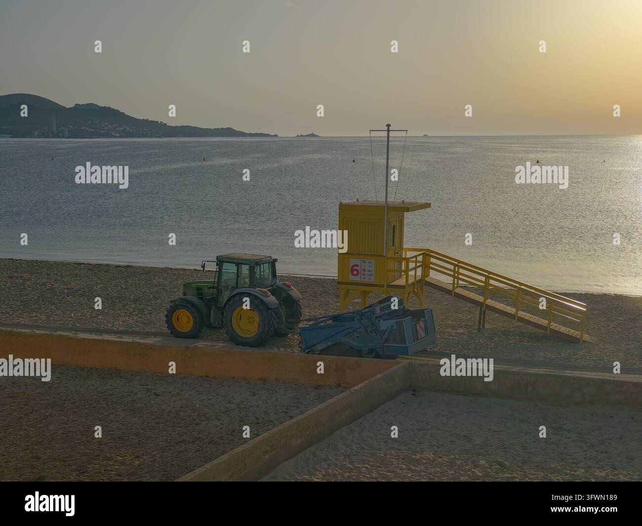 Un trattore verde con ruote gialle tira una macchina per la pulizia della spiaggia su una spiaggia sabbiosa accanto a una torretta di salvataggio gialla brillante. Il mare calmo si estende fino a Foto Stock