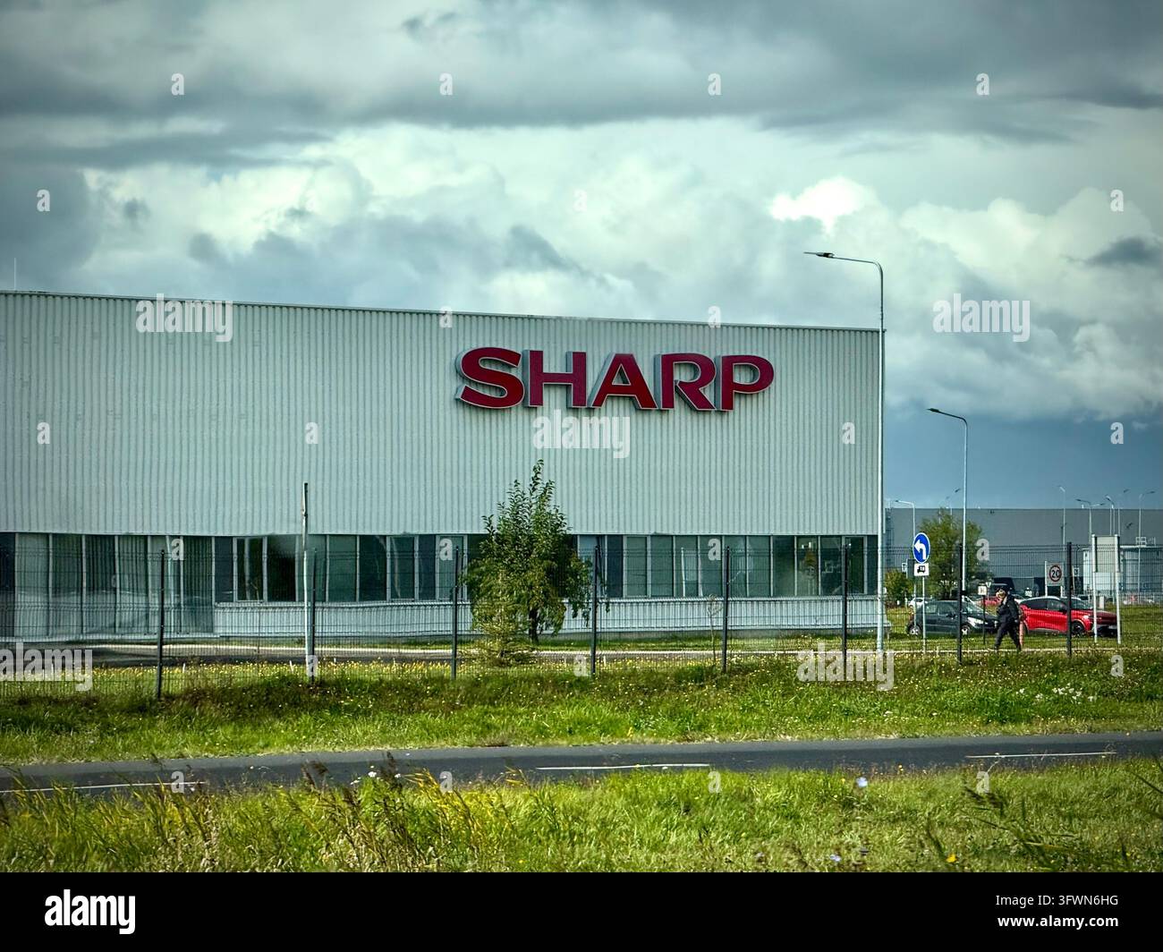 un moderno edificio industriale grigio con un grande logo rosso "SHARP" sulla facciata sotto un cielo drammatico e nuvoloso. - Immagine stock catturata con smartphone
