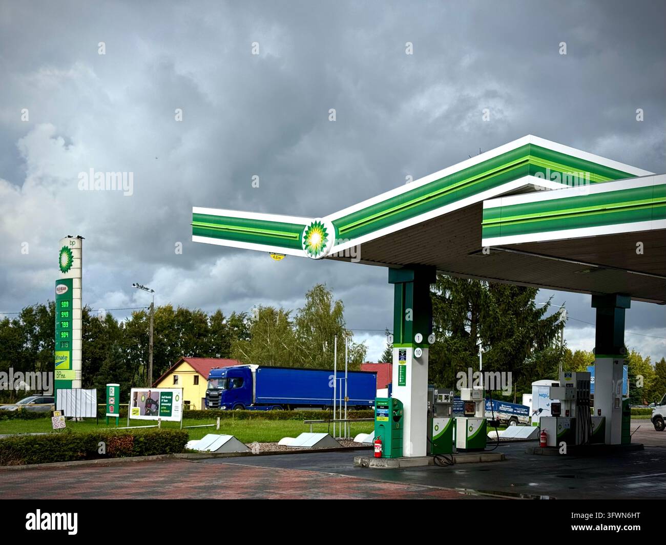 Stazione di servizio BP con il suo caratteristico tetto verde e bianco, erogatori di carburante e colonna totem dei prezzi sotto un cielo cupo e drammatico. - Immagine stock catturata con smartphone