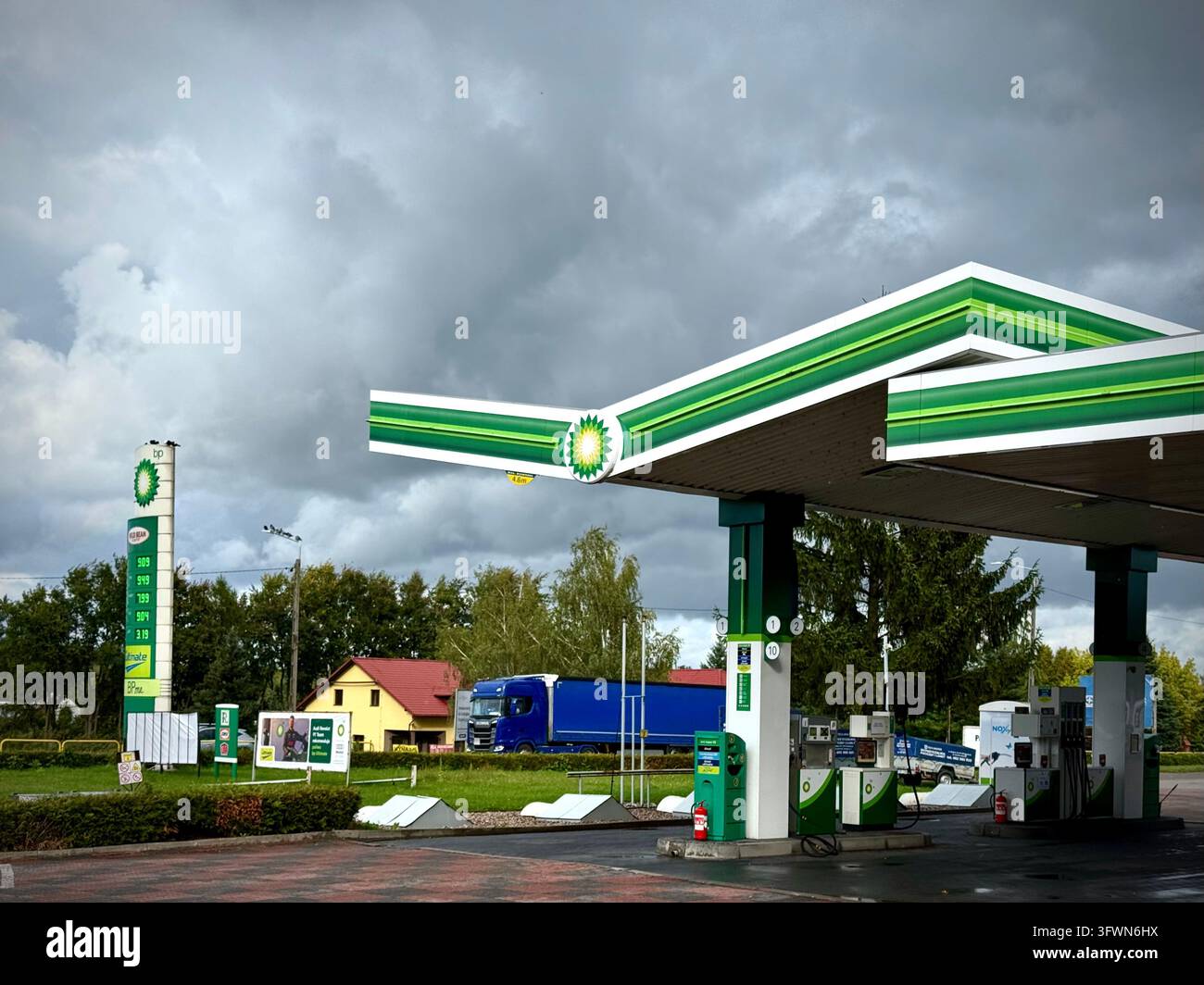 Stazione di servizio BP con il suo caratteristico tetto verde e bianco, erogatori di carburante e colonna totem dei prezzi sotto un cielo cupo e drammatico. - Immagine stock catturata con smartphone