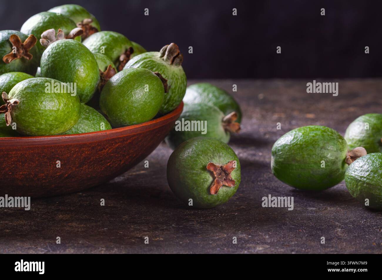 Colata studio di cibo all'altezza degli occhi che mostra feijoas verdi in una ciotola di argilla rustica su un tavolo scuro e texture, illuminato lateralmente. Foto Stock