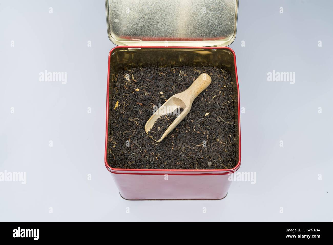 Foglie di tè nero sfuse in una scatola metallica rettangolare aperta con un piccolo misurino di legno, isolate su sfondo chiaro Foto Stock