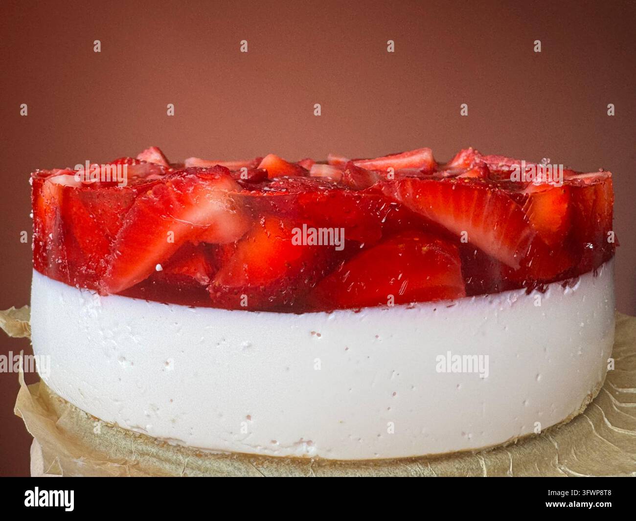 Vista laterale di una cheesecake senza cottura con uno strato di crema bianca liscio ricoperto di fragole fresche a fette disposte in una glassa rossa. Il dolce poggia su un biscotto - Immagine stock catturata con smartphone