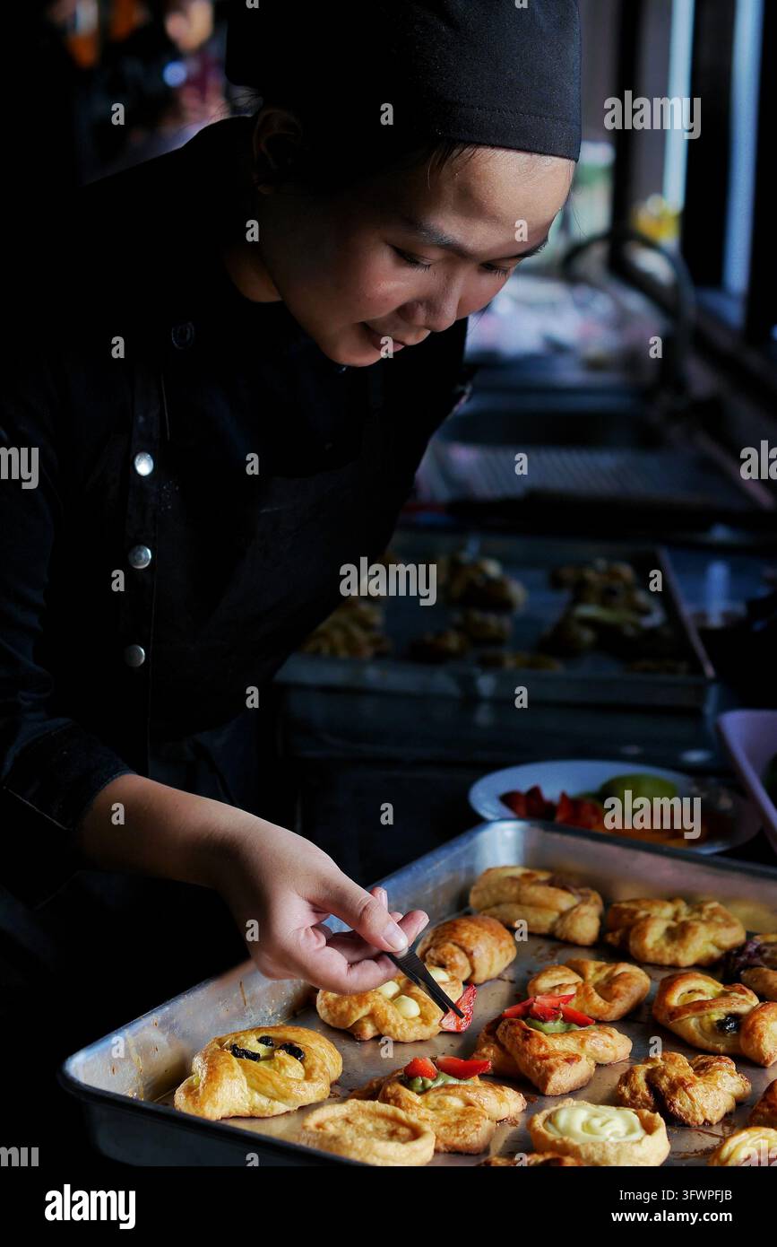 Una pasticcera in abiti neri decora vari dolci o pane cotto utilizzando pezzi di frutta su una teglia da forno. Foto Stock