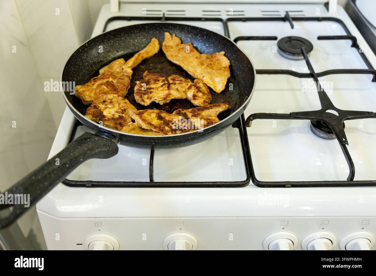 Filetti di pollo dorati fritti cuociono in una padella nera su un fornello a gas bianco, mostrando una semplice preparazione di pasti fatti in casa in una cucina domestica. Foto Stock