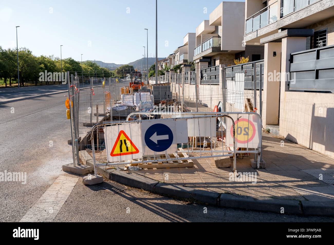 Barriere temporanee e segnali stradali segnano lavori di manutenzione stradale accanto a case residenziali moderne, con un'area del marciapiede scavata e infrastrutture di servizio Foto Stock