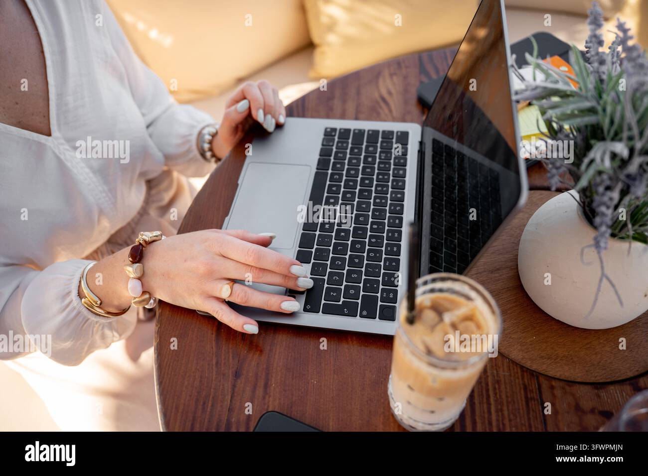 Primo piano di una donna d'affari che utilizza un laptop a un tavolo di un caffè sul mare, con caffè freddo e piante, rappresentante il lavoro remoto, il freelance e il dig Foto Stock
