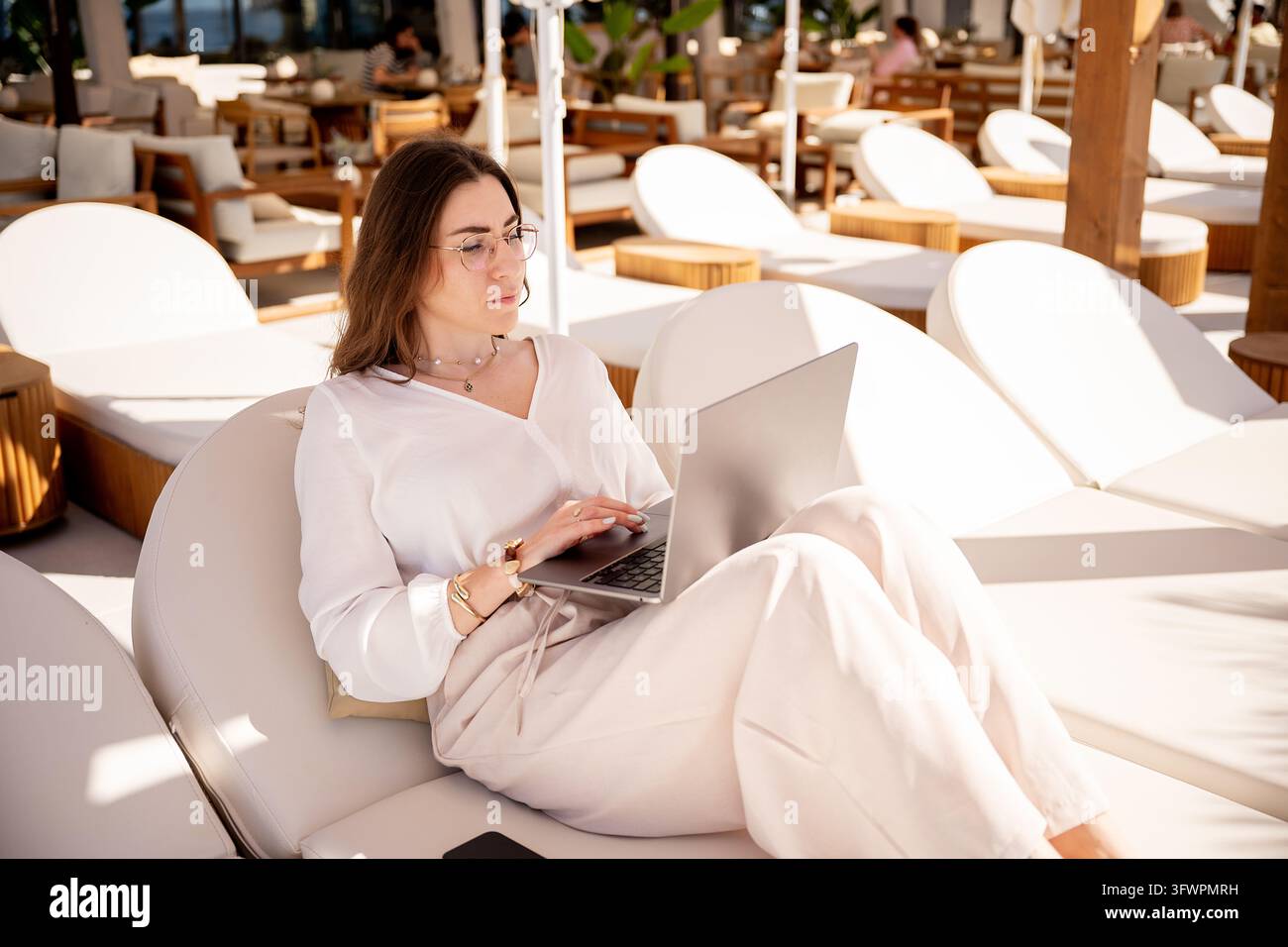 Donna d'affari in abiti bianchi lavora da remoto su un laptop mentre si rilassa in un caffè di lusso sul mare, circondata da sedie a sdraio illuminate dal sole e da un'atmosfera calorosa Foto Stock