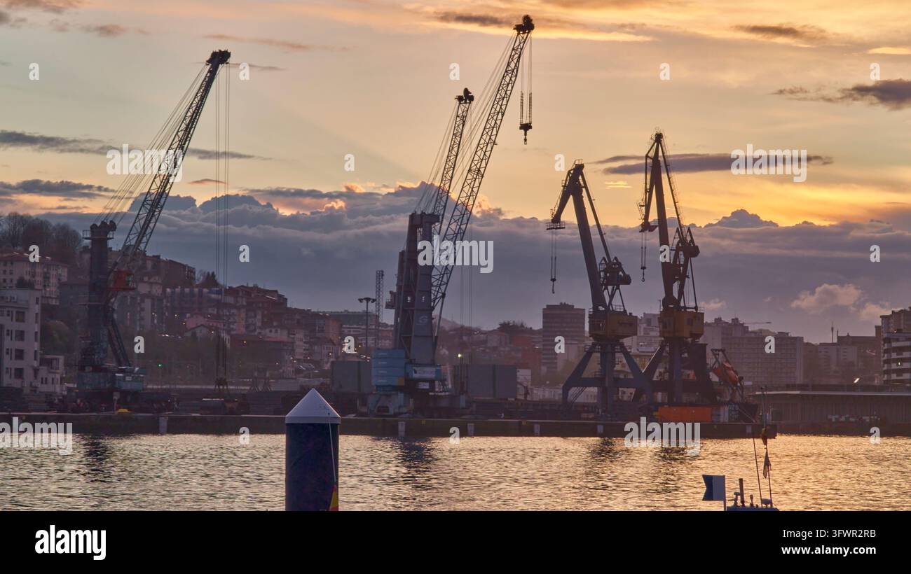 Pasai Donibane, Spagna – 9 marzo 2026: Gru portuali industriali al tramonto, logistica marittima e infrastrutture commerciali globali con silhouette del porto merci al crepuscolo, terminale costiero e navigazione Foto Stock