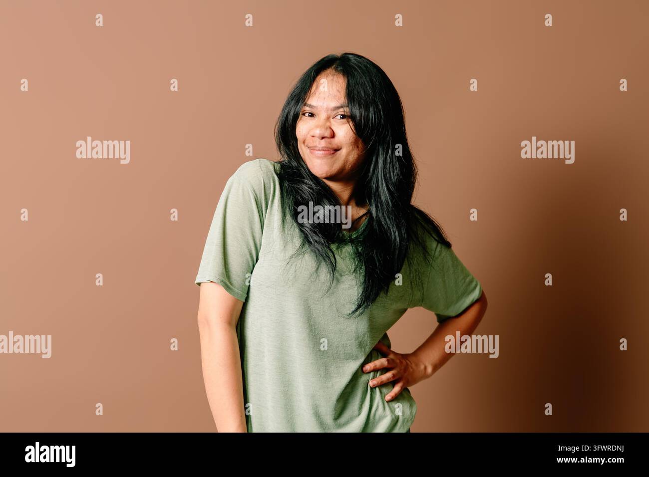 Donna asiatica sorridente con maglietta verde chiaro che alza le spalle con un sorriso carino e malizioso su uno sfondo studio marrone. Foto Stock