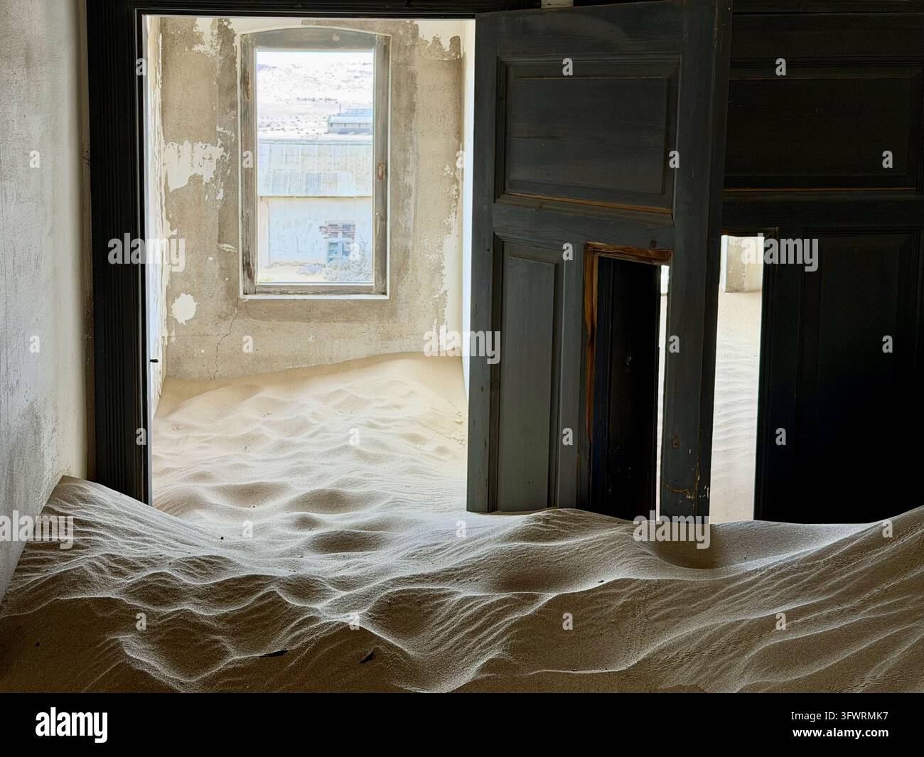 Porte di legno scuro si ergono sopra cumuli di sabbia pallida a forma d'onda all'interno di un edificio abbandonato di Kolmanskop in Namibia. - Immagine stock catturata con smartphone