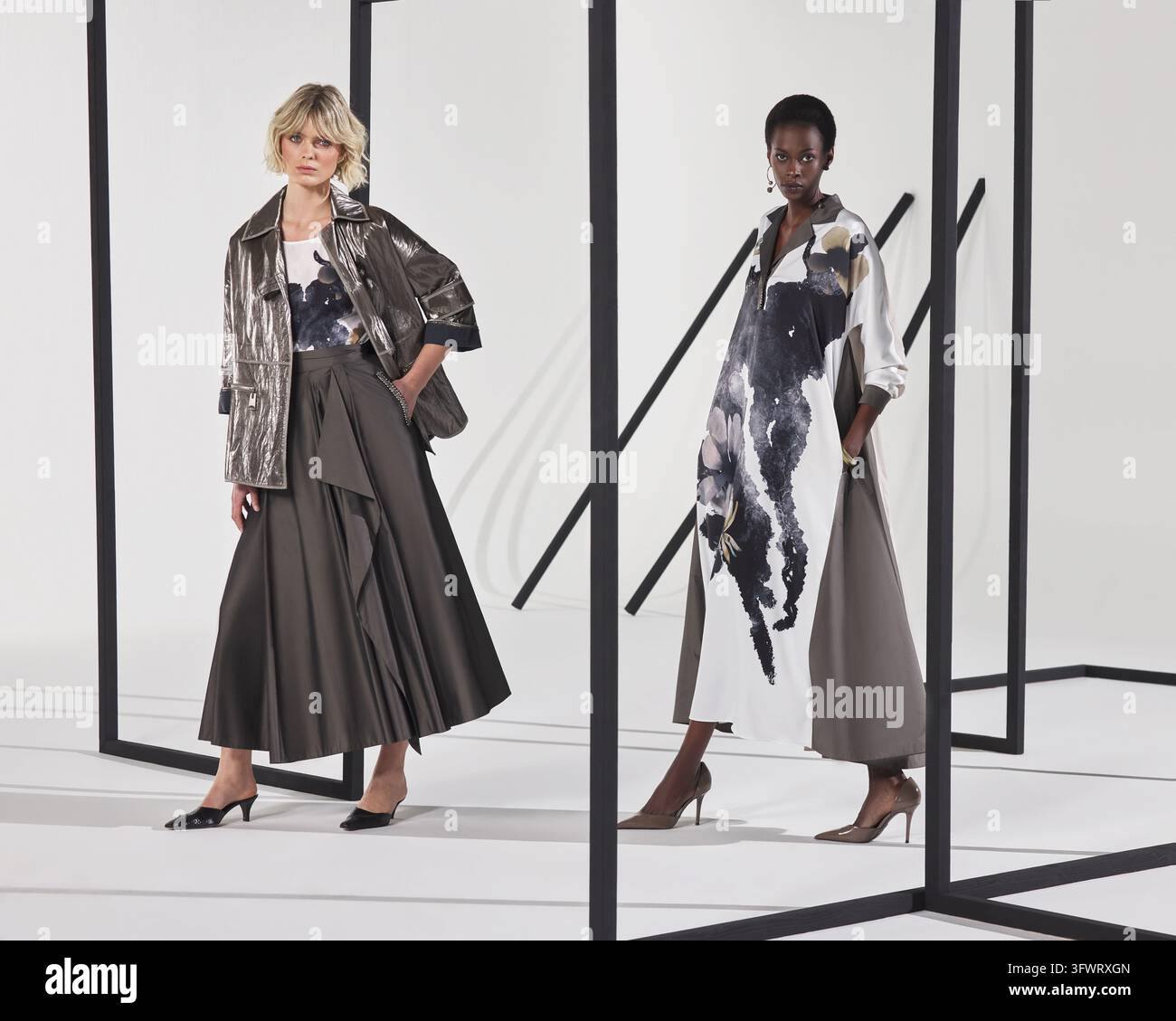Due modelle in un set di studio, una con una giacca di pelle metallica e una gonna lunga scura, l'altra con un lungo vestito bianco con stampa acquerello. Foto Stock