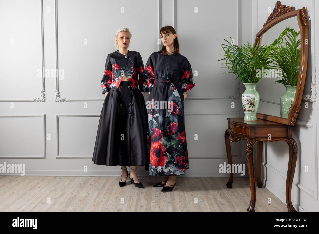 Inquadratura ampia di due modelli in nero con stampe di rose rosse, uno con una giacca floreale corta e una gonna nera ampia, l'altro con un abito lungo a stampa di rose. Foto Stock