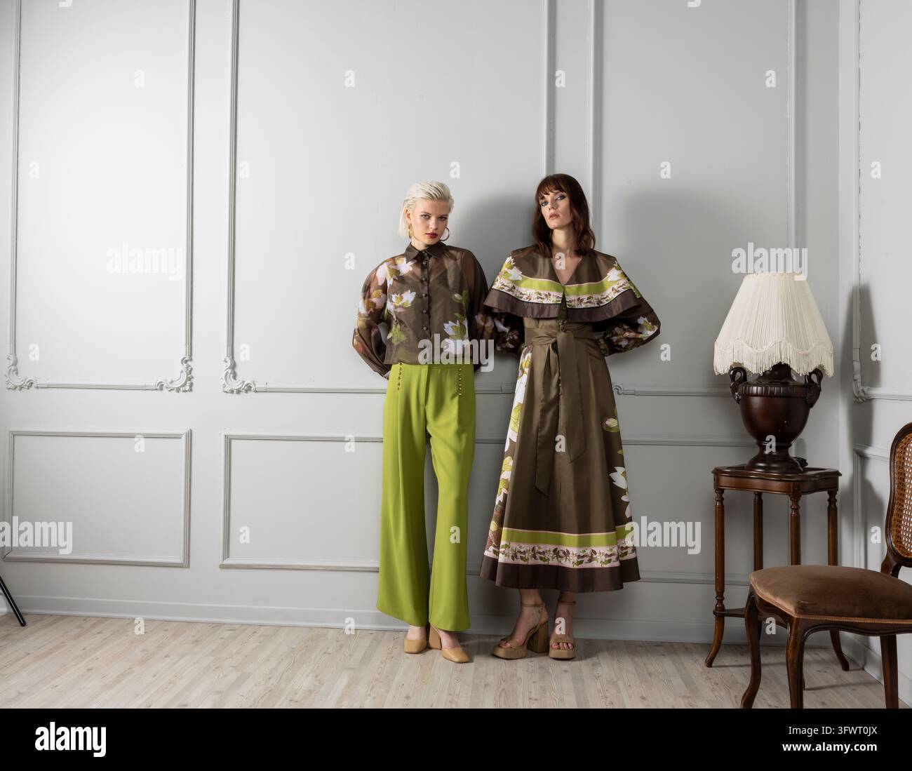 Inquadratura ampia di due modelli, uno con una camicetta marrone floreale trasparente e pantaloni verde lime, l'altro con un vestito lungo in raso oliva con un colletto a capelet stampato. Foto Stock