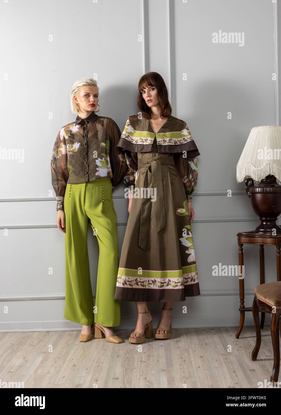 Foto a tutta lunghezza di due modelli, uno con una camicetta trasparente a stampa magnolia e pantaloni verde lime, l'altro con un vestito di raso olivastro e un mantello stampato. Foto Stock