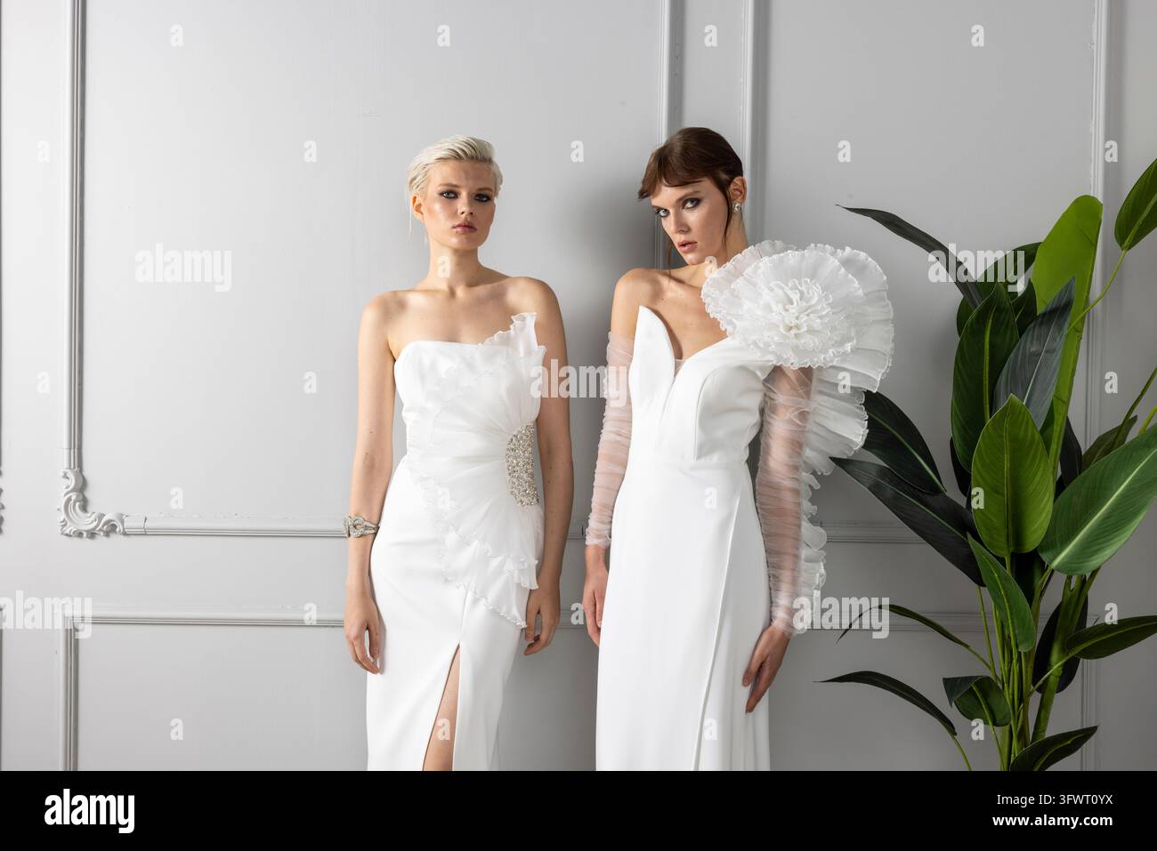 Inquadratura a tre quarti di due modelli in bianco, un abito senza spalline con vita in cristallo e un abito da sera a scollo a V con una spalla a balza scultorea in organza. Foto Stock