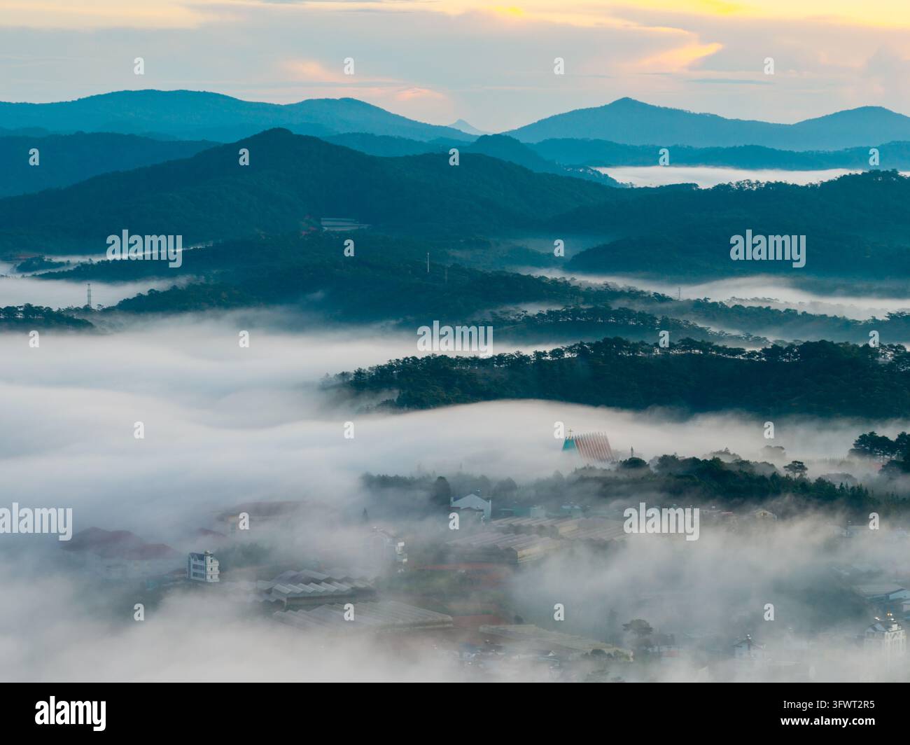 Vista aerea delle montagne nebbiose e delle foreste di pini che circondano la città di Da Lat, con edifici che emergono attraverso la fitta nebbia mattutina a Xuan Huon Foto Stock