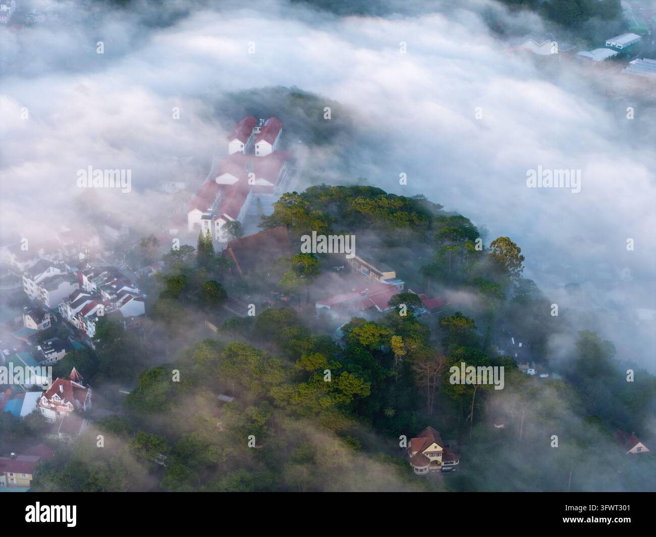Vista aerea di edifici con tetti rossi incastonati nelle foreste di pini parzialmente coperti da una fitta nebbia mattutina a Da Lat, Lam Dong, Vietnam. Foto Stock