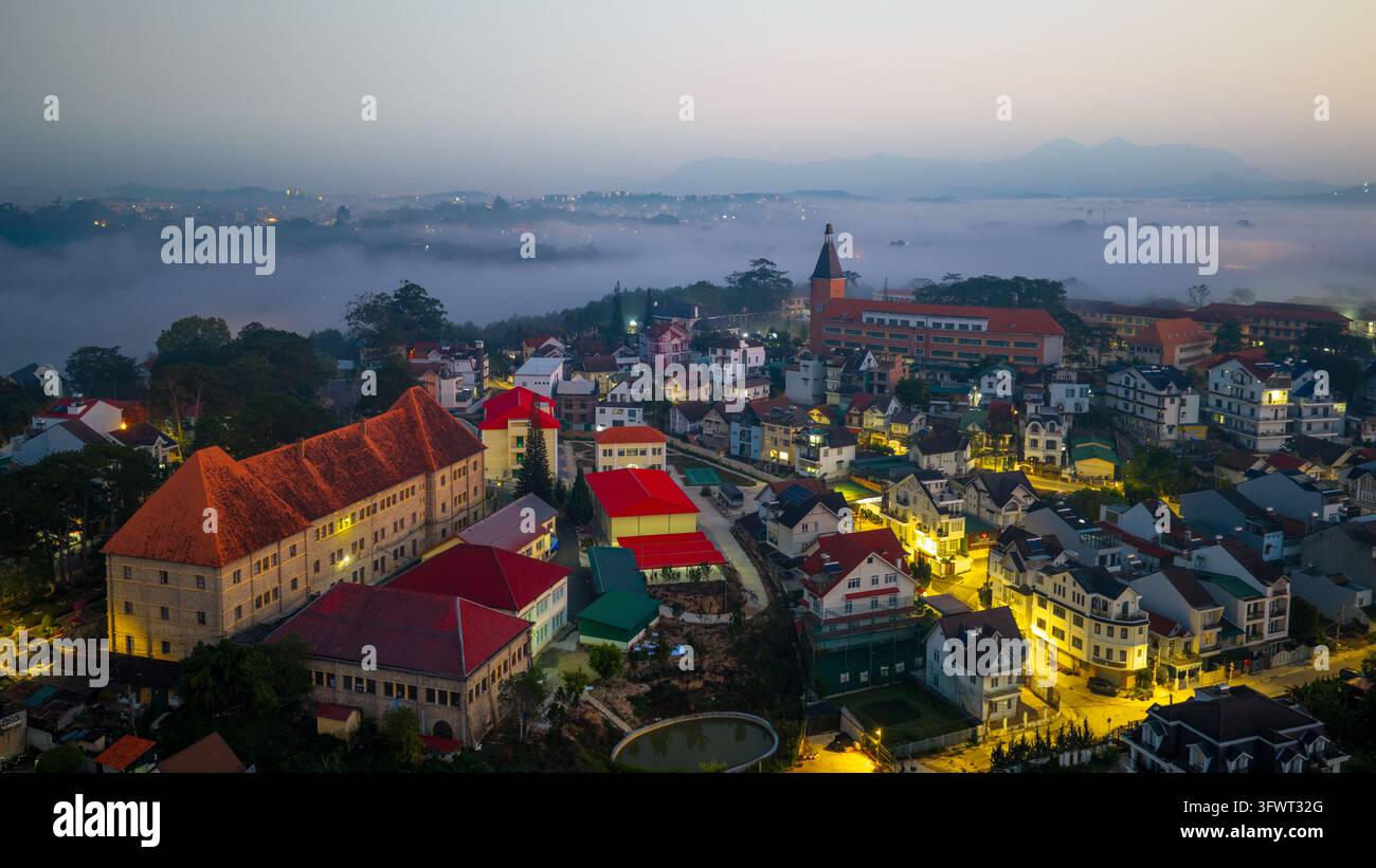 Vista aerea del Collegio Pedagogico e delle case colorate circostanti nella nebbia mattutina, con colline ondulate e paesaggio urbano a Da Lat, Lam Dong Foto Stock