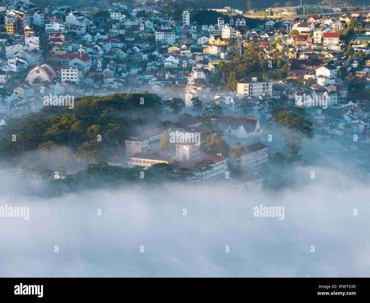 Vista aerea della città di Da Lat incastonata tra foreste di pini e colline ondulate, con una fitta nebbia mattutina che copre la valle e tetti colorati visibili Foto Stock