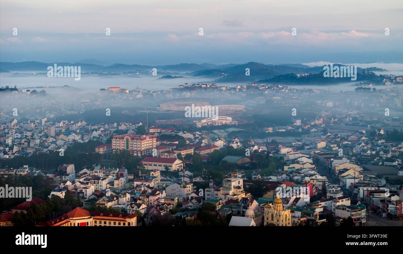 Vista aerea della città di Da Lat incastonata tra colline ondulate coperte dalla nebbia mattutina con un'architettura urbana densa, tetti colorati e uno stadio a Da L Foto Stock