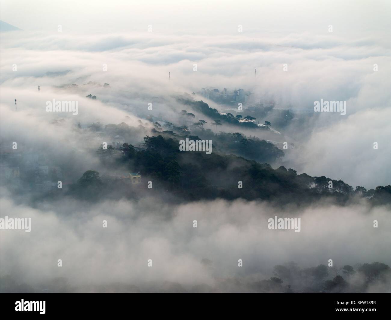 Vista aerea della città di Da Lat parzialmente oscurata da una fitta nebbia mattutina e nuvole, con colline ricoperte di pini e strutture urbane che emergono attraverso la foschia Foto Stock