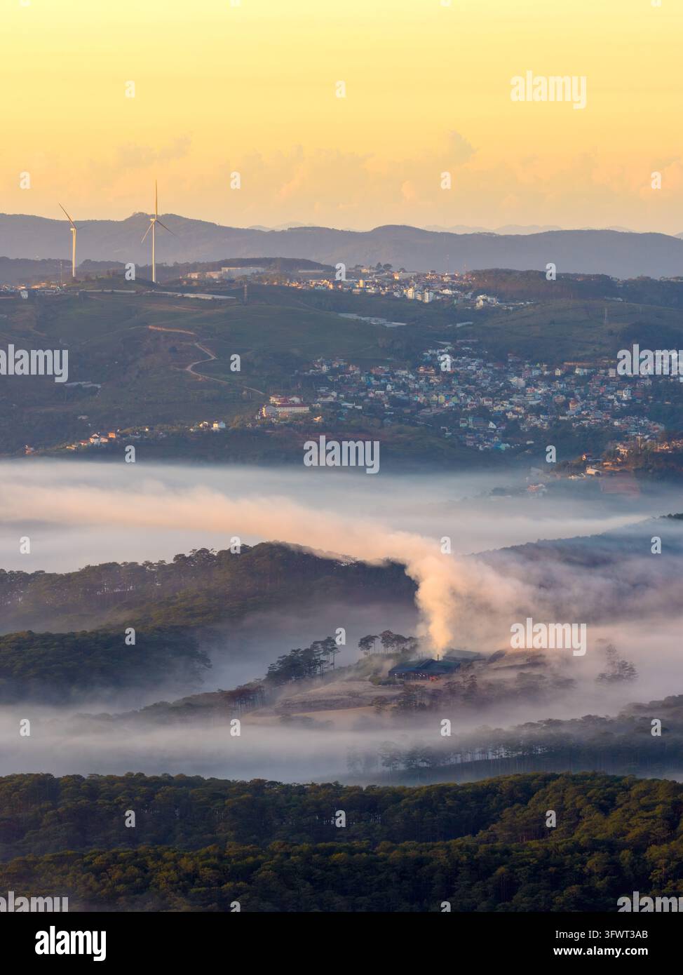 Vista aerea del paesaggio nebbioso di Da Lat all'alba, con colline ondulate, turbine eoliche all'orizzonte e un piccolo insediamento che emerge dal t Foto Stock