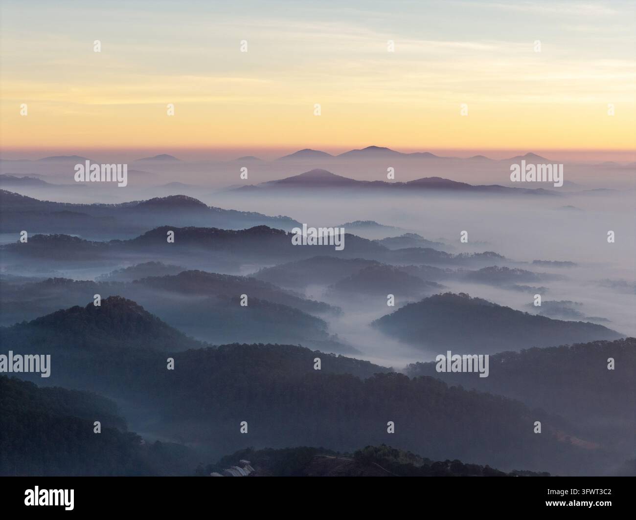 Vista aerea di montagne nebbiose e colline stratificate all'alba con una luce dorata soffusa sulle valli a Da Lat, Lam Dong, Vietnam. Foto Stock