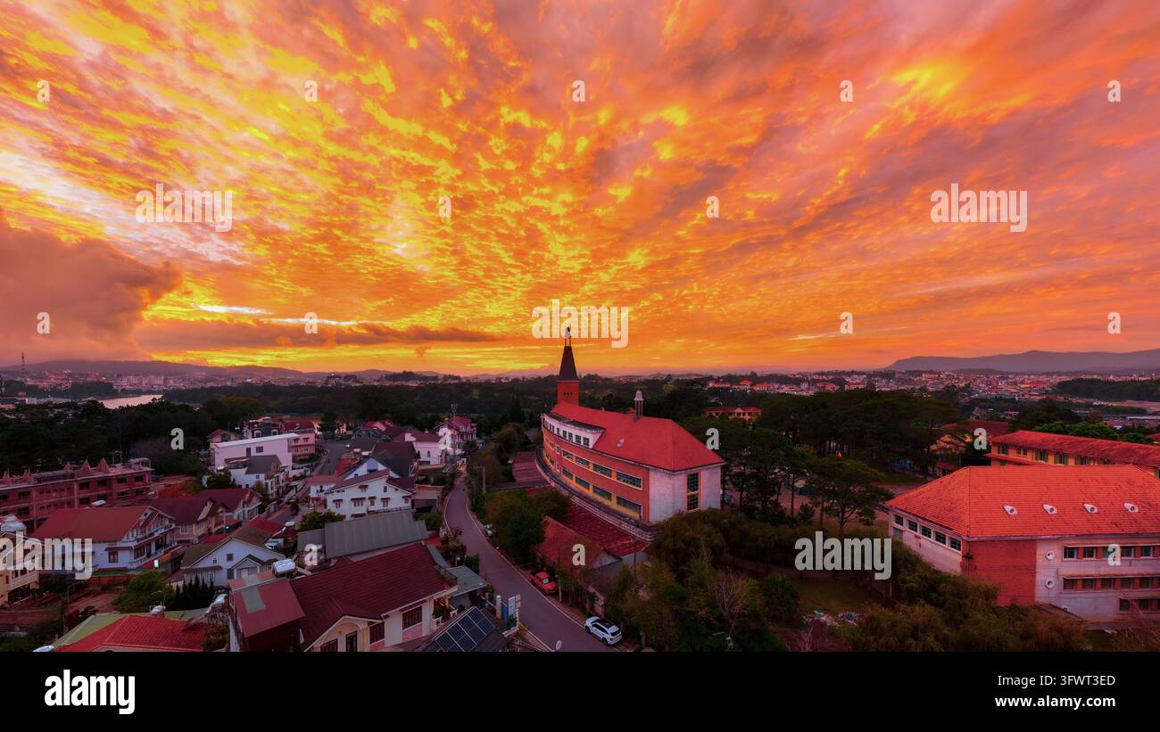 Vista aerea del Collegio Pedagogico di Da Lat con il suo iconico campanile, circondato da case con tetti rossi e una vegetazione lussureggiante sotto un vivace arancione Foto Stock