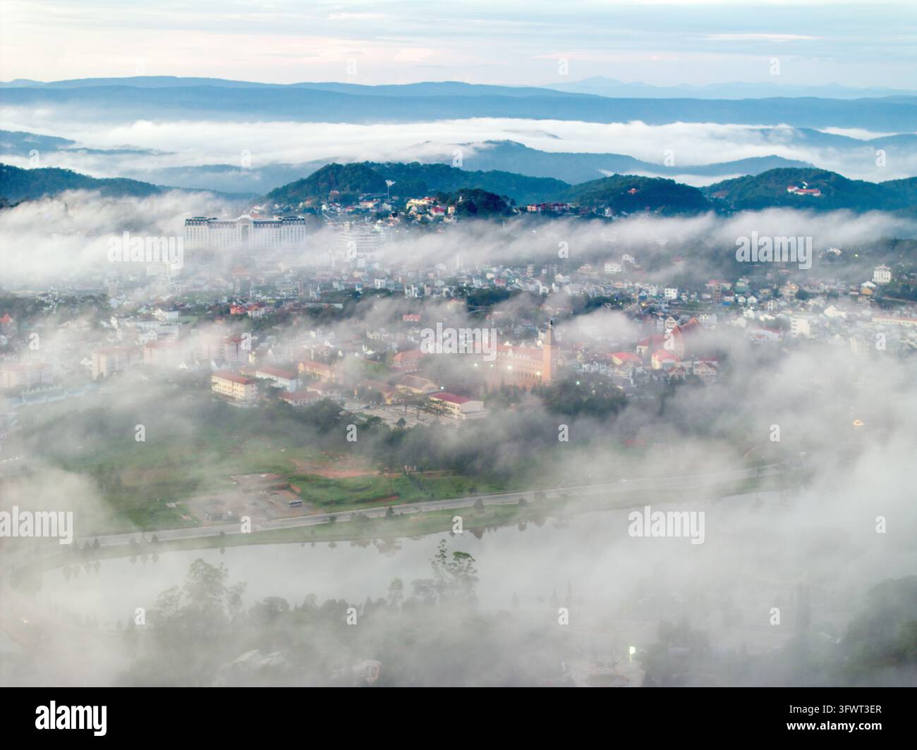 Vista aerea del lago Xuan Huong avvolto nella nebbia mattutina con le colline circostanti e gli edifici urbani a Da Lat, Lam Dong, Vietnam. Foto Stock
