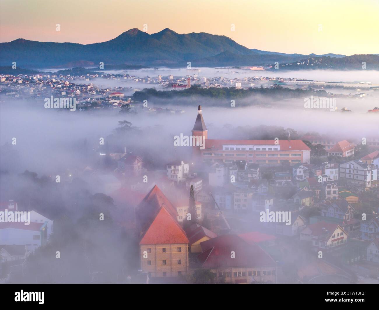 Vista aerea della città nebbiosa con i suoi edifici iconici dal tetto rosso, il campanile della chiesa e le montagne circostanti all'alba a Da Lat, Lam Dong, Vietnam. Foto Stock