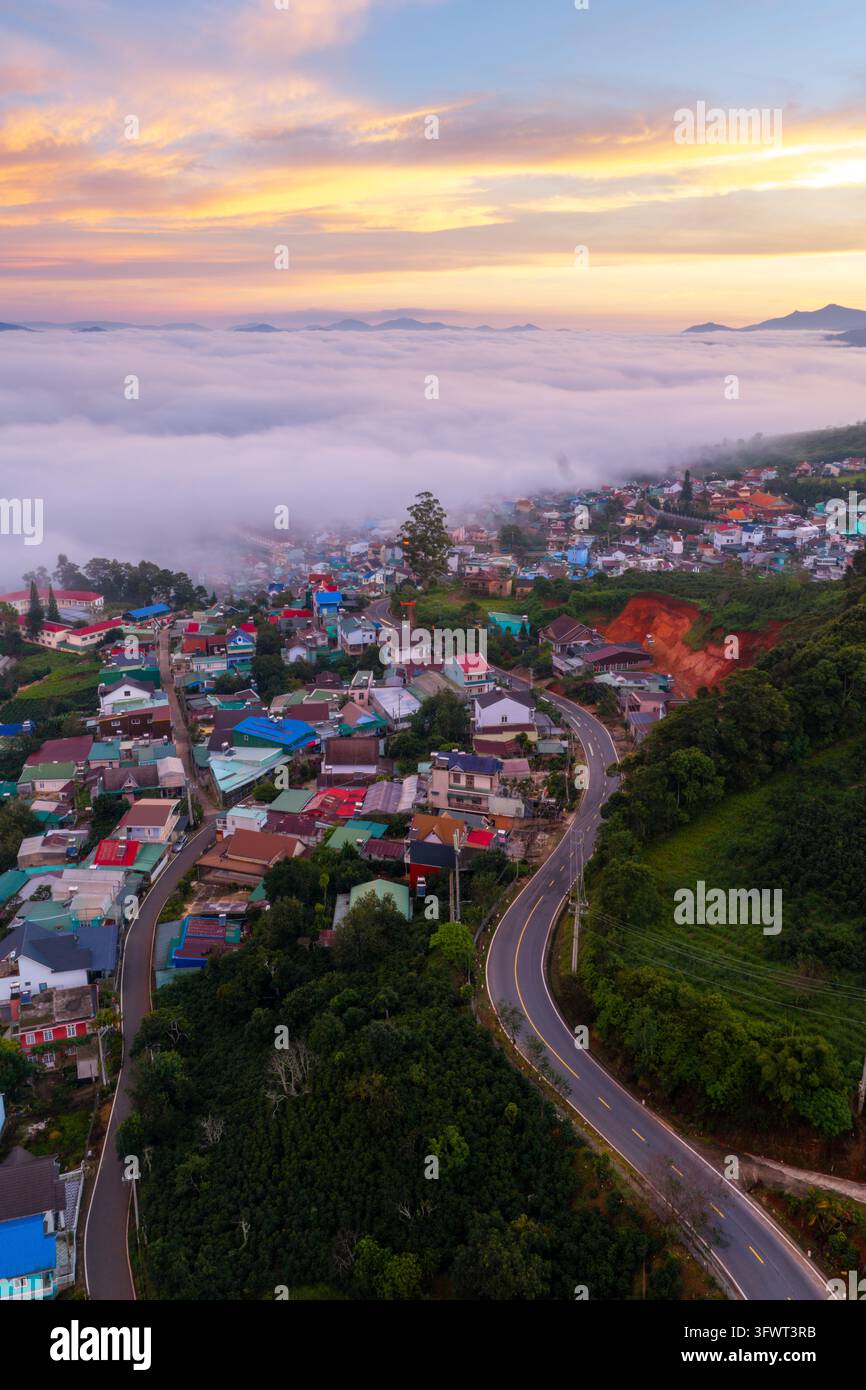 Vista aerea della città di Da Lat annidata tra le colline, con una strada tortuosa che passa attraverso case colorate sotto un cielo vivace al tramonto sopra le nuvole a Da Lat Foto Stock