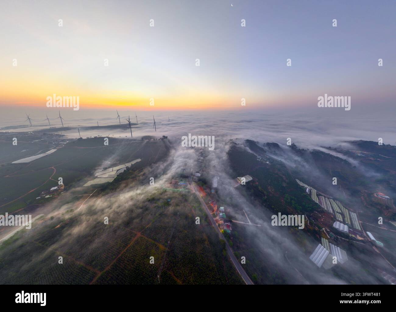 Vista aerea di turbine eoliche che si ergono su un mare di nuvole sopra le colline ondulate e le serre di Da Lat all'alba. Da Lat, Lam Dong, Vietnam. Foto Stock