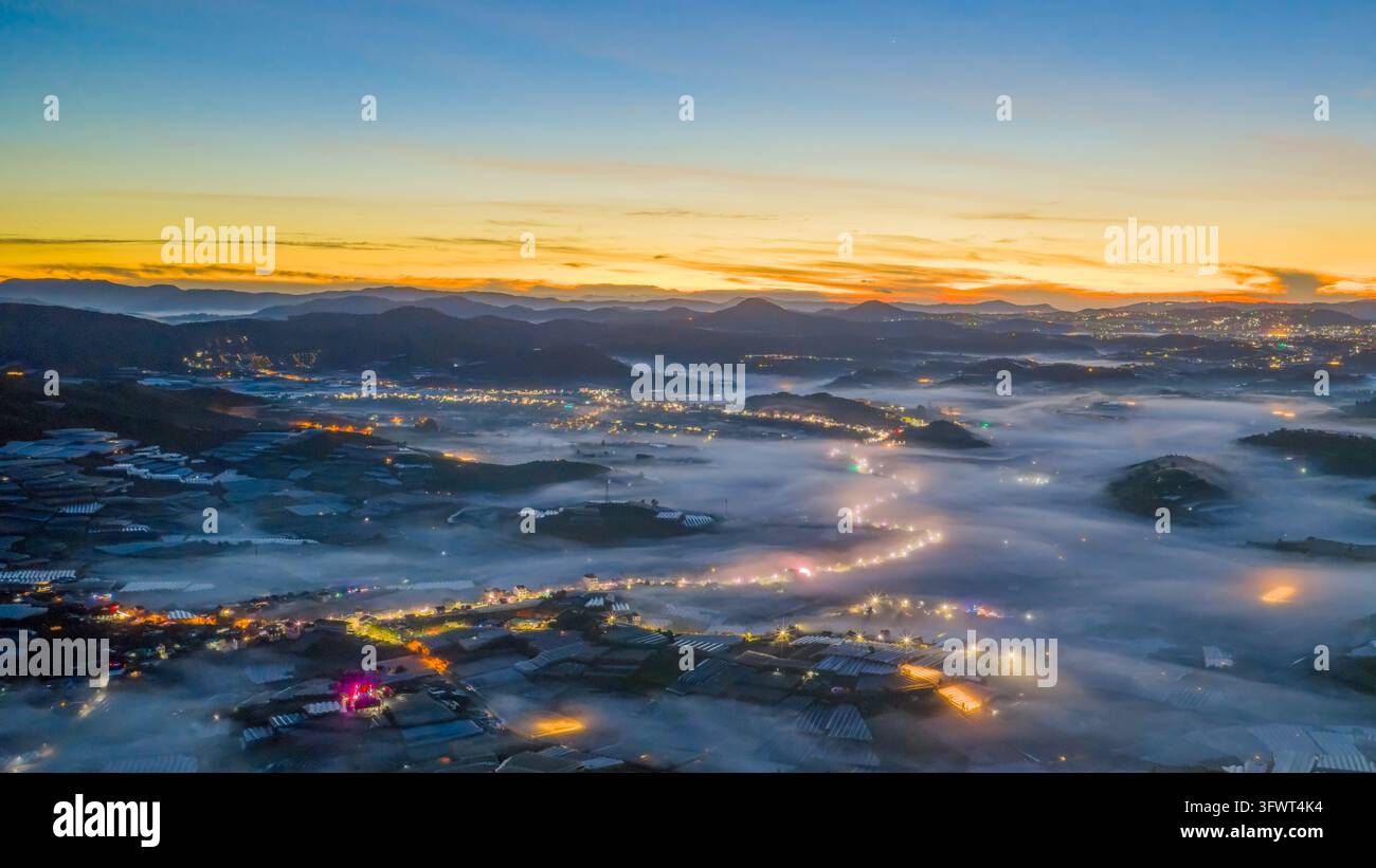 Vista aerea del paesaggio nebbioso di Da Lat all'alba, con luci brillanti di serre che illuminano la valle coperta di nebbia nelle terre alte di Da Lat, L Foto Stock