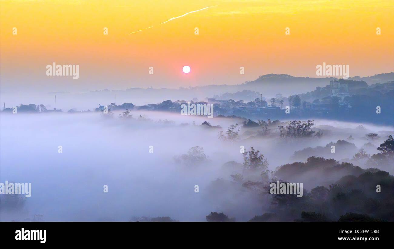Vista aerea del paesaggio nebbioso all'alba, con una fitta nebbia che copre la valle, alberi sparsi e edifici lontani sotto un cielo dorato a Da Lat Foto Stock