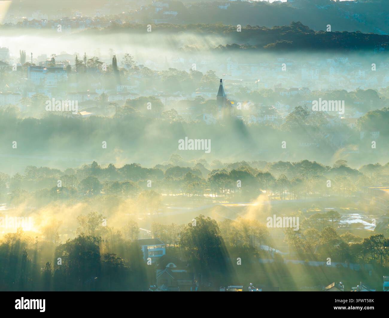 Veduta aerea di un paesaggio nebbioso con raggi di sole che filtrano attraverso alberi ed edifici nella luce del primo mattino a Da Lat, Lam Dong, Vietnam. Foto Stock