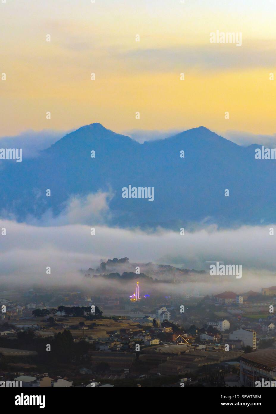 Vista aerea della città annidata tra le montagne con nebbia bassa e un edificio landmark luminoso all'alba, Da Lat, Lam Dong, Vietnam. Foto Stock