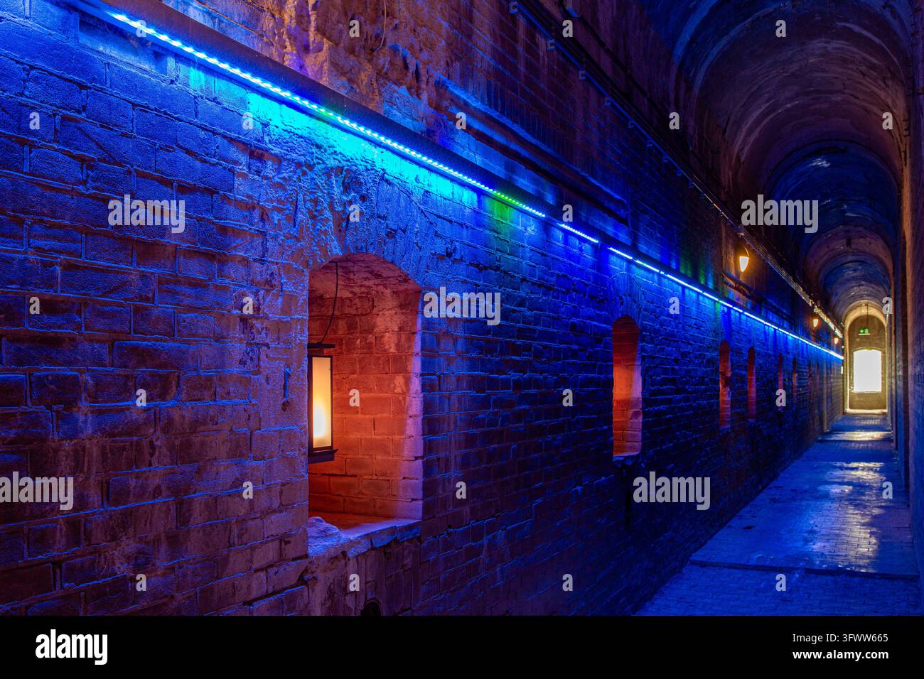 Vista del corridoio storico in mattoni del Fort Pampus, illuminato da moderne strisce LED blu e lanterne calde nelle nicchie delle pareti, Gooise Meren, Nort Foto Stock