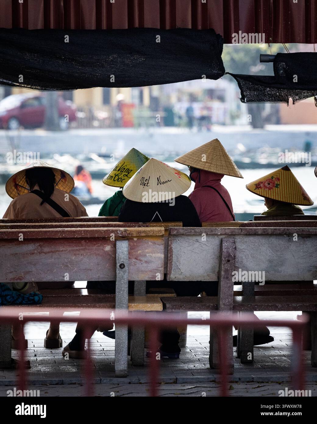 Ninh Binh, Vietnam - 8 gennaio 2026: Un gruppo di persone che indossano tradizionali cappelli a cono vietnamiti seduti su panchine vicino all'acqua. Foto Stock