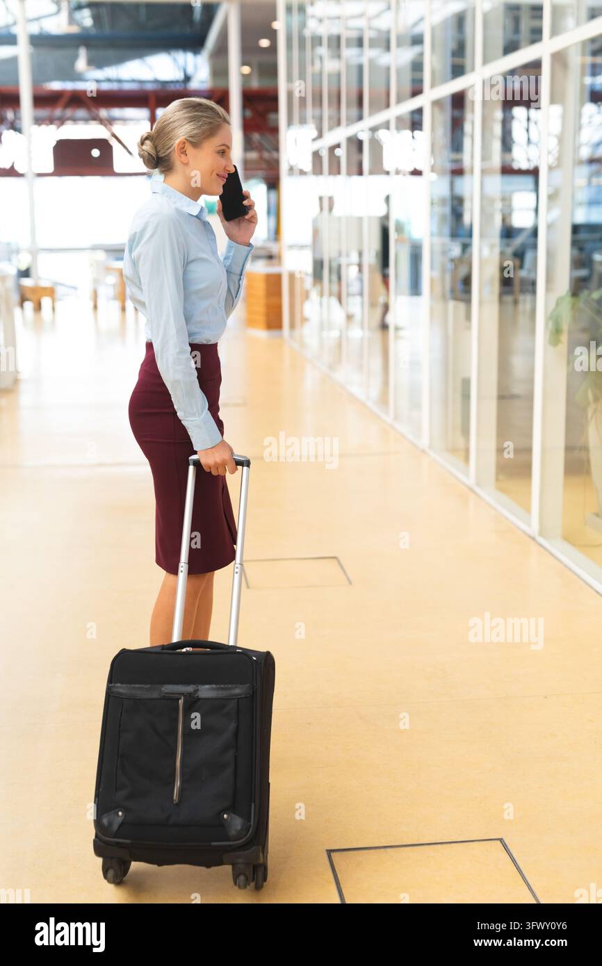 Vista laterale di una donna d'affari caucasica che parla al telefono cellulare mentre è in piedi con il bagaglio in un corridoio dell'ufficio. Business aziendale internazionale e diversificato Foto Stock