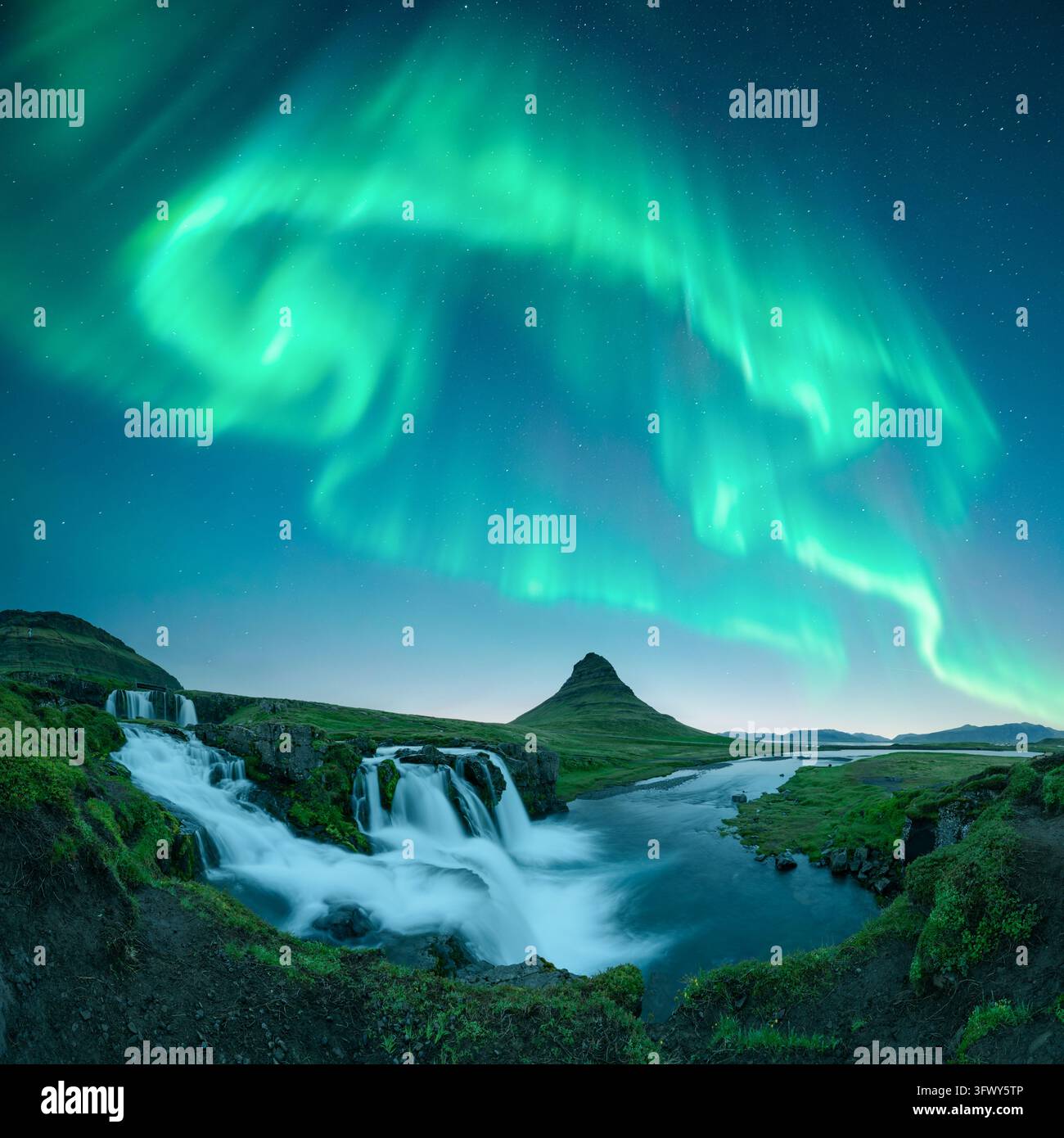 L'aurora boreale di un verde vivace brilla sopra la cascata di Kirkjufellsfoss e il vulcano Kirkjufell. L'erba rigogliosa e l'acqua che scorre creano un paesaggio islandese drammatico. Le aurore boreali in Islanda Foto Stock